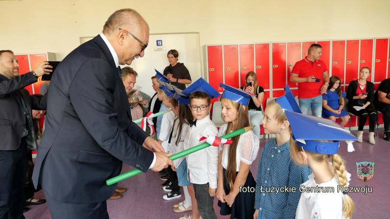 Mężczyzna w garniturze opiera duży ołówek na ramieniu jednego z kilku dzieci w szeregu. Mężczyzna ma okulary, a Dzieci są ubrane w stroje galowe i czapeczki akademickie.